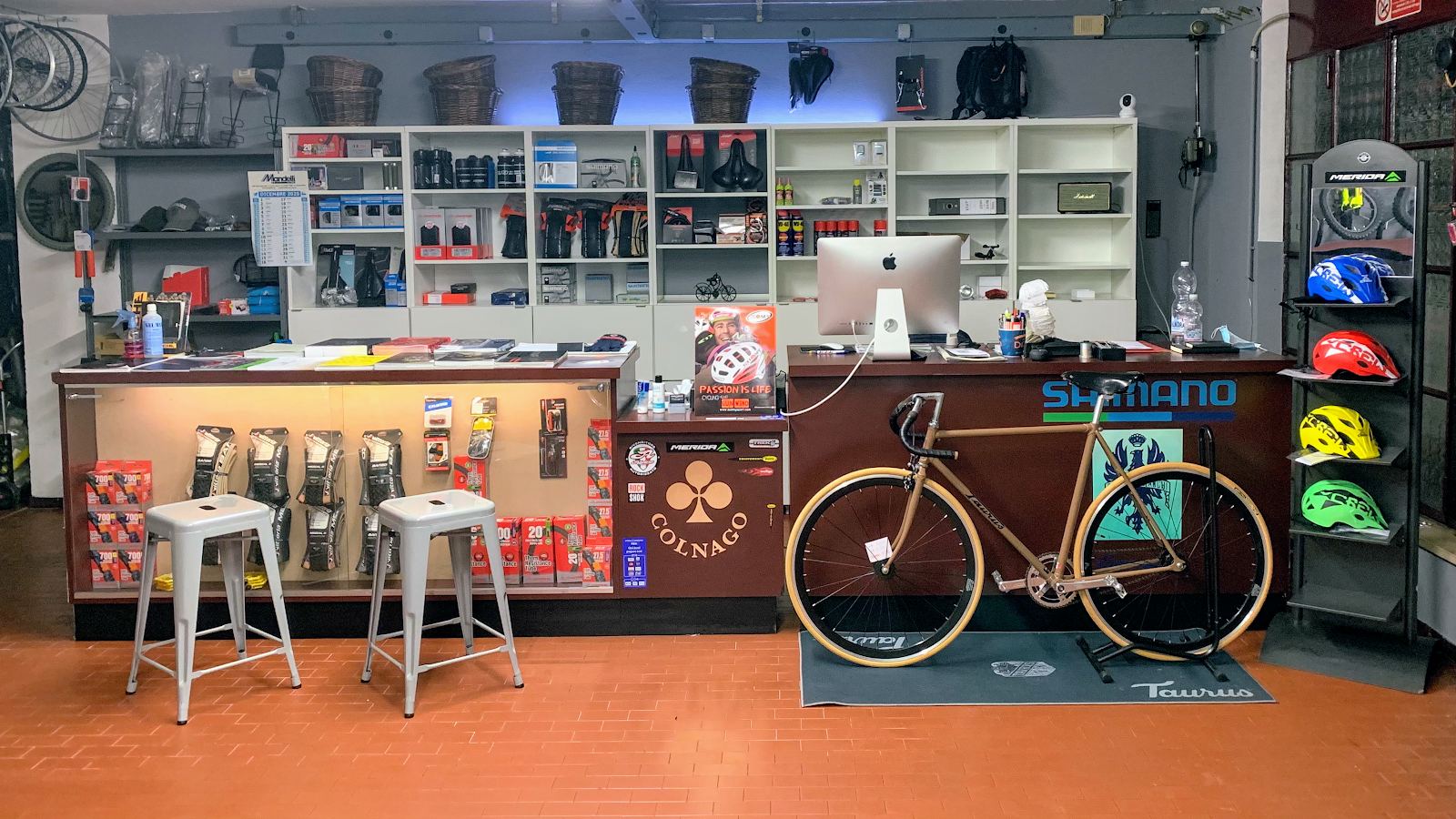 Il banco del negozio: copertoni e camere d'aria in vetrina, scaffali con selle e ricambi, una bici da pista color bronzo e una fila di caschi colorati