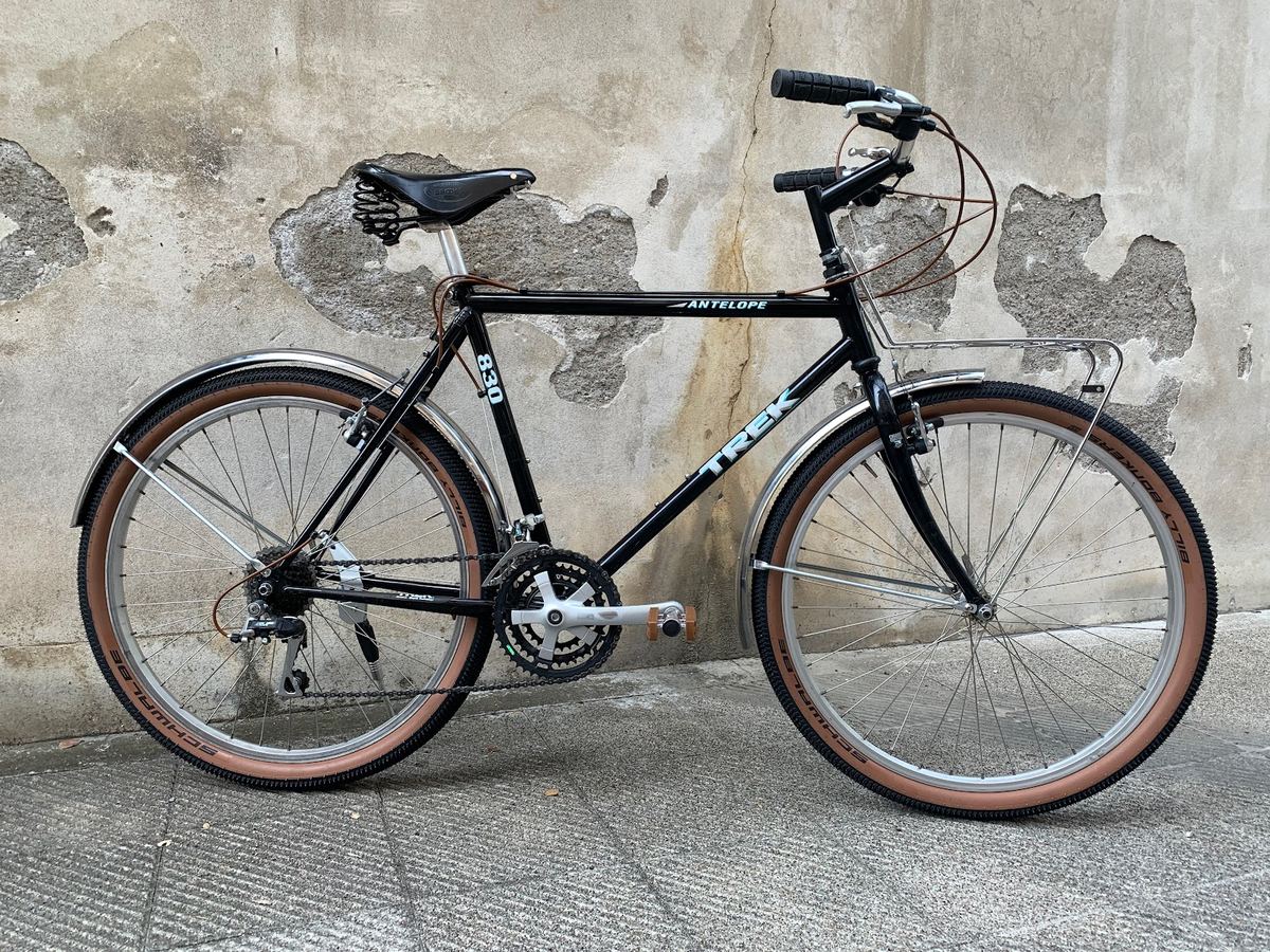Bici Trek Antelope nera con sella in cuoio e portapacchi anteriore, appoggiata a un muro